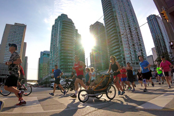 A large group of runners crossing Queen's Quay at Yonge, the sun setting dramatically between condo towers behind them